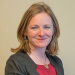 Freelance science communication team member Allanah Robinson of GoodSense - smiling to camera wearing a dark jacket, red top and with mid length mid brown hair