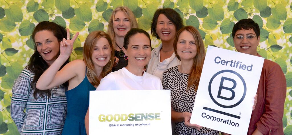 B Corp Certified Sustainability marketing & ethical communications team GoodSense celebrate becoming BCorp - seven people stand infront of a leafy green background smiling and holding signs. The signs are a GoodSense logo and a Certified B Corporation logo. One person is making a peace sign.