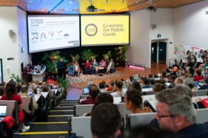 Room full of people watching a panel of speakers on stage below two images one reads Economy for Public Good