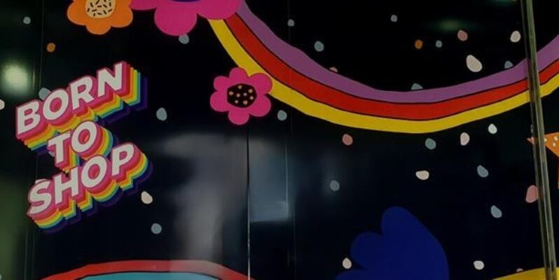 Consumerism - Photograph of mural on shopping mall wall with the text Born to Shop and fraphics of stars, flowers and circles in flouro bright colours on a black background